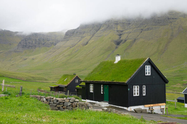 Scenic shot of a traditional Scandinavian village in the vast green valley on a cloudy day in Faroe Islands. Cool black houses with white window frames and grassy roofs in the untouched countryside.