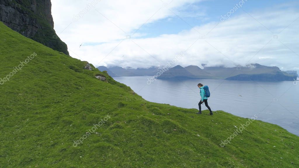 AERIAL: Female tourist hikes up a grassy hill with a perfect view of the ocean on a cold morning. Young woman on hiking trip walks up a steep mountain overlooking the vast sea and the green mainland.