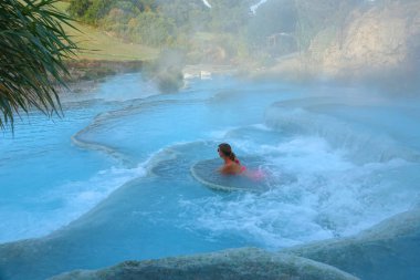 Genç beyaz bir kadın beyaz bir taş çıkıntıya canlandırıcı termal hamam İtalya içinde dayanıyor. Kız pembe bikini Toskana sıcak doğal havuzlarda rahatlatıcı. Kadın turist puslu açık kaplıcada zevk.