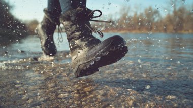 CLOSE UP, LOW ANGLE, DOF: Energetic young woman in fuzzy black boots playing by the tranquil stream on a lovely autumn day. Unrecognizable playful hiker girl running and splashing pure river water.