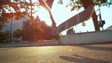 LOW ANGLE, LENS FLARE, CLOSE UP: Unrecognizable young man flips the skateboard with his feet. Cool skater riding and doing tricks in the city on a sunny autumn day. Unknown skateboarder doing flips.
