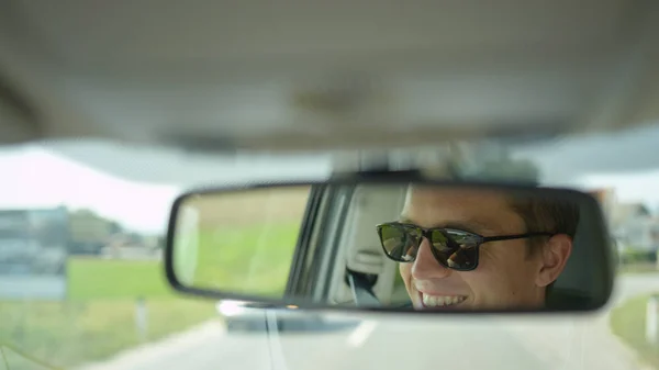 CLOSE UP, DOF: Young Caucasian male smiles while driving his car down the country road on his way to work. Cool shot of the reflection of a cheerful male driver in the rear view mirror of his car.