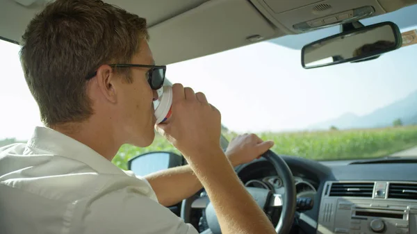 クローズ アップ 若い白人男性は 美しい晴れた日に空の田舎道を運転中コーヒーを飲みます ハンサムな男が運転する道路の旅行中に茶色の紙カップから熱いお茶の Sip を取る — ストック写真