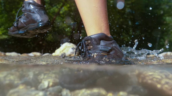 HALF UNDERWATER, CLOSE UP, DOF: Unrecognizable active woman hiking on a perfect sunny day steps out of glassy river water. Young female trekker in waterproof leather boots walking along the stream.