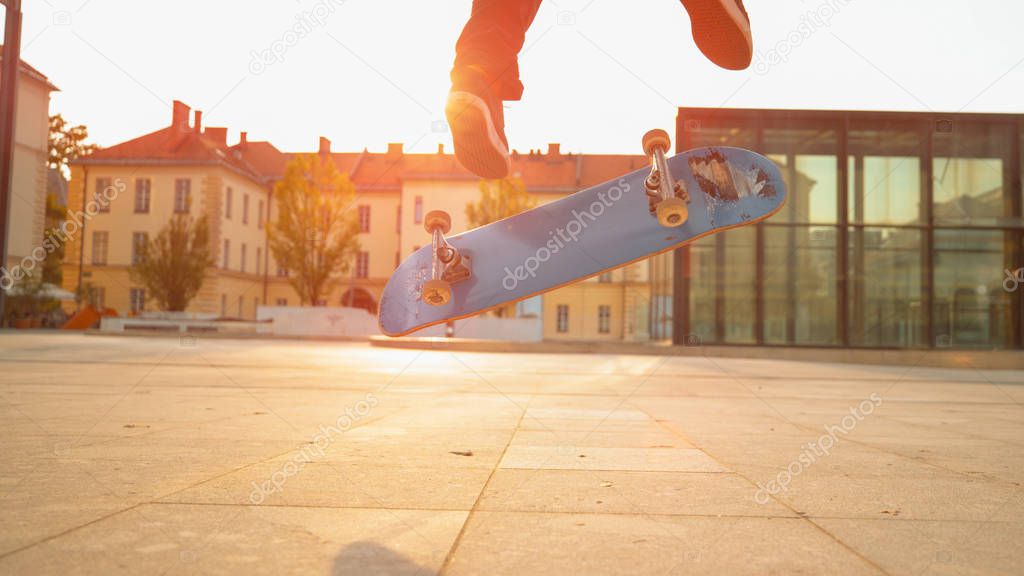 LOW ANGLE, CLOSE UP, LENS FLARE: Unrecognizable athletic skateboarder doing a difficult trick on an idyllic sunny evening. Cool shot of a blue skateboard flipping under unknown athletic man's feet.