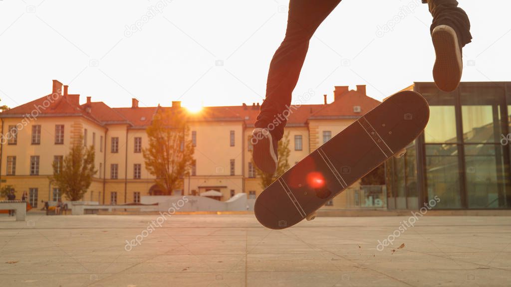 LENS FLARE, CLOSE UP, LOW ANGLE: Young skater lands a cool trick while in skateboaring in the urban park at idyllic sunset. Male skateboarder does a fakie during an evening session at a city square.