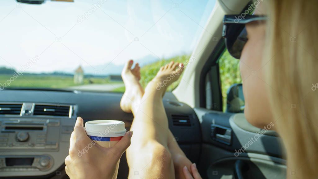 CLOSE UP: Young Caucasian woman resting her feet on the dashboard holds a cup of takeaway coffee during a scenic summer road trip through the countryside. Happy blonde girl enjoying a cup of coffee.