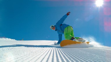 Düşük açılı, Lens Flare: kış güneşte taze bakımlı bir yamaç oyma neşeli genç erkek snowboard. Güneşli Alpler'de sürme sırasında serin serin bir elin yaptığından beyaz snowboard görüntüsünü sürükleyin.