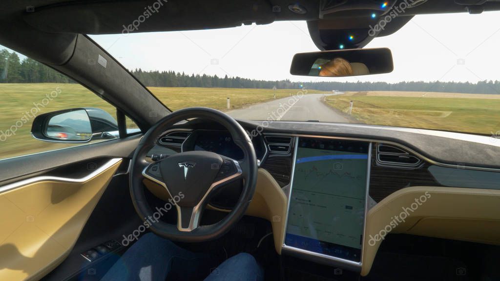 TESLA AUTONOMOUS CAR, March 2018 - POV: Young woman cruises down an empty country road in a self steering Tesla car. Sunny rural landscape surrounds the autonomous vehicle driving down the road.