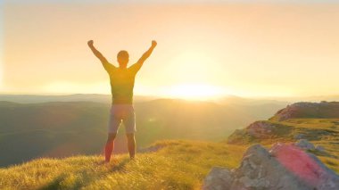 Dron, Lens Flare, kopya alanı: Parlatıcı güzel yaz günbatımı görmek için kollarını başının üzerine çimenli tepeye hiking sonra yetiştirme muzaffer erkek uzun yürüyüşe çıkan kimse üzerinde parlak akşam güneş ışınları. Heyecanlı atlet