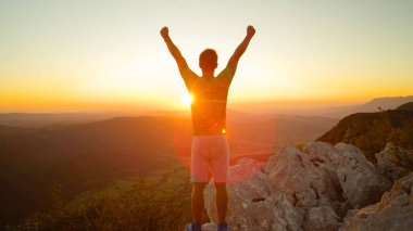 CLOSE UP, LENS FLARE: Happy hiker raises his arms overhead after seeing the sunset from the top of the big mountain. Unrecognizable male tourist celebrates reaching top of the hill on a sunny morning.