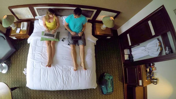 TOP DOWN: Young Caucasian couple on vacation in Vietnam working on their laptops while sitting on the king size bed. Female and male tourists surfing the internet from their comfortable hotel room.