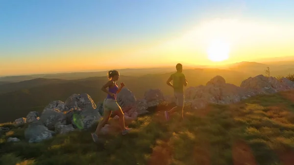 ドローン 太陽フレア 夕暮れ山道に沿って実行しているスポーティな観光カップルの後ろに飛んでいます 黄金の朝太陽光線をあてる運動の若い男性と女性の困難落ちたレースを実行するためのトレーニング — ストック写真