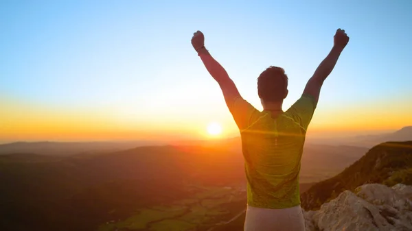 CLOSE UP, LENS FLARE, COPY SPACE: Cheerful male hiker celebrates reaching the summit at idyllic sunrise. Unrecognizable sporty man outstretches arms as he observes the scenic green valley below him.