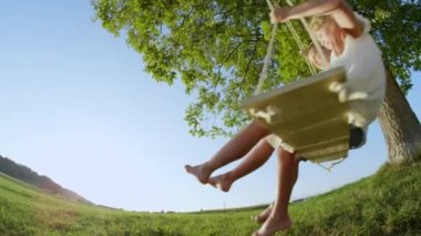 Yavaş Hareket Düşük Açı: Yaz aylarında büyük yeşil bir ağacın altında tahta bir salıncak üzerinde sallanan birbirlerine tutan genç çocuklar çift. Sıcak bir günde erkek ve kız kucaklaşıyor. Sarılmak kardeşler yalınayak sallanan.