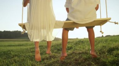 Slow Motion Close Up: Güneşli akşamlarda büyük ahşap salıncakta oturan tanınmayan çocuklar. Yalınayak küçük erkek ve kız kardeşi güneşli yaz sallanan. Romantik akşamerkek ve kız arasında genç aşk