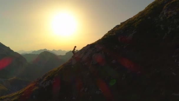 空中剪影 在金色的夏日夜晚 在女性徒步旅行者旁边飞行 使她在崎岖不平的山坡上飞翔 年轻女子徒步旅行者爬上陡峭的山在金色的日落在风景如画的山 — 图库视频影像