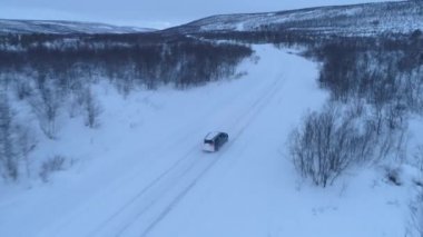 Aerial Close Up: Kaygan karlı yolda, karla kaplı dağa doğru çıplak ağaçların yanından geçen arabanın üzerinde uçmak. Lapland, Finlandiya'da yolculuk yapanlar. Oto uzak kış vahşi ile seyahat