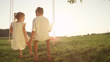 Slow Motion Close Up: Küçük kız eğlenceli sıçrayan ve altın günbatımında ahşap salıncak üzerinde oturan genç tatlı çocuk ile el ele tutuşurken şarkı. Aşık çift sıcak bir yaz akşamı eğleniyor.