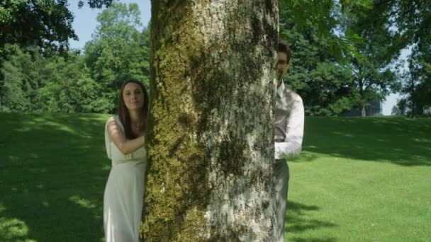 スローモーションポートレート陽気なカップルは 木の後ろから覗き カメラに微笑みます 恋に落ちた男女は 公園の真ん中に設置された木の幹で遊び心のあるポーズをとります 夫と妻は屋外で時間を楽しんで — ストック動画