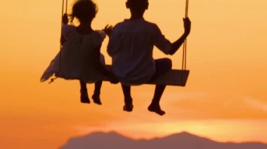 Yavaş Hareket Silueti, Close Up Genç çift altın ışık salıncak üzerinde sallanan ise gün batımı bakıyor. Tanınmayan çocuklar muhteşem gün batımında sallanıyorlar. Küçük erkek ve kız kardeş altın gün batımının tadını çıkarıyor