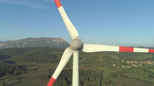 空中クローズアップ 緑の田園地帯で白い金属の刃を照らす春の日差し 強風で回転する風力発電機 近くの村の再生可能な電力源を表す — ストック動画