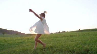 Slow Motion Close Up Silly kız dans ve geç öğleden sonra yeşil bir çayır üzerinde dönen. Akan beyaz bir elbiseiçinde dönen goofy çocuk. Genç kız kardeş çimenli bir alanda yalınayak dans.