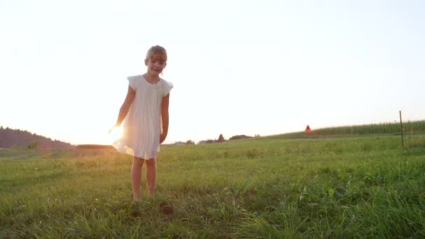 スローモーションクローズアップ白いドレスで愚かな小さな女の子は 午後遅くに屋外で楽しみを持っています 日没時に草原で遊んで夏を楽しんでいるグーフィーの子供 興奮した妹が元気に拍手 — ストック動画