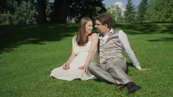 スローモーション 若いカップルは 美しい日当たりの良い公園で草の上に座って遊び心にキス 暖かい晴れた日に両親が優しさを分かち合う 夏に屋外で素晴らしい時間を過ごしている新婚夫婦 — ストック動画