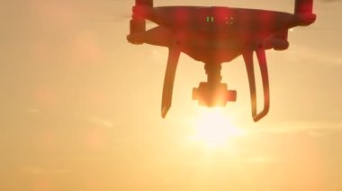 Yakın Çekim Lens Flare Siluet Küçük filme drone altın günbatımı gökyüzü üzerinde uçan. Altın ışıkta çekim eklenmiş kamera ile beyaz quadcopter drone. Gündoğumu gökyüzünde uçan Quadrocopter helikopter