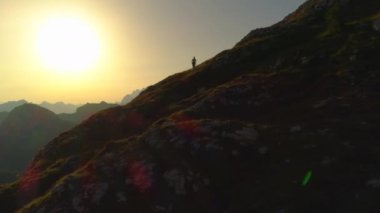Hava Siluet kopya alanı: tepe boyunca yürüyen ve güneşli dağ da yukarı doğru dönen kadın yürüyüşçü yanında uçan. Genç kadın güzel yanmış turuncu günbatımı nda dik kayalık yamaca kadar trekking