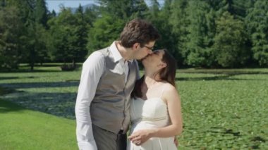 SLOW MOTION CLOSE UP: Loving husband with his arms wrapped around his wife, gives her a tender kiss. Devoted young couple embrace and smile at the camera. Man tenderly caresses wife's pregnant belly.