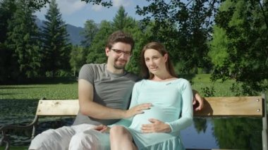 Portre: Güzel genç çift sevinçle ahşap bankta oturan ve kameraya gülümseyerek bekliyor. Gururlu gelecek anne parkta güzel bir güneşli gün zevk ve yakında çocuk doğacak okşama.