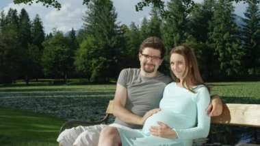 yavaş hareket portre: güzel Kafkas çift yeşil parkta romantik bir bankta sarılma. Gülümseyen genç kadın ve adam güzel bir güneşli günde beklenen annenin hamile bebeğini okşuyor.