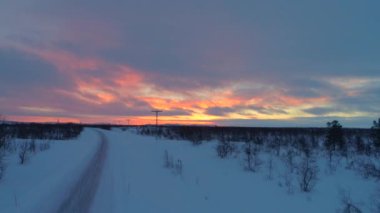 Hava: Karlı kış vahşi doğada çıplak ağaçların yanından geçen boş otobanın üzerinde uçmak, Lapland, Finlandiya. Dramatik gün doğumunda manzaralı yol. Güzel turuncu günbatımında wintry kırsal buzlu yol