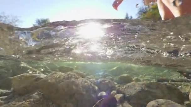 ハーフハーフ レンズフレア 緑の峡谷を走る川を渡るハイカーのための良いグリップを提供する茶色の革の登山ブーツ 冷たい川を踏む女性は 澄んだ水の中に波紋と泡を作成します — ストック動画