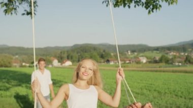 SLOW MOTION CLOSE UP: Joyful young woman is pushed on the swing by happy young man. Girl's golden hair is flowing and gets messy in the summer air as she swings on the rope swing in a sunny meadow.
