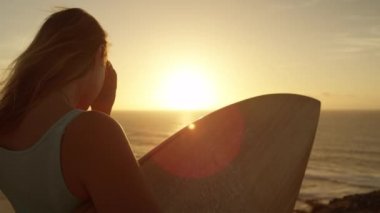yavaş hareket lens parlama: tanınmaz genç sörfçü kız gün batımı ve dalgalar bakarak saçları ile oynar. Bir sörf tahtası tutan, uçurumda duran kadın gün batımında hafif yaz rüzgarında saçlarını ayarlar.