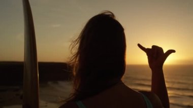 Slow Motion Close Up: Uzun akan saçları ile tanınmayan kız altın gün batımısama shaka işareti veren bakıyor. Kadın sörfçü eliyle gevşek sinyal asmak yapıyor. Hawaii el hareketi kullanan kadın
