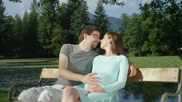 肖像画 晴れた日に木製のベンチに座って 最初の子供が抱き合い 優しくキスをすることを期待する若いカップル 緑の自然の中でキスを期待する白人カップルの美しいロマンチックなショット — ストック動画