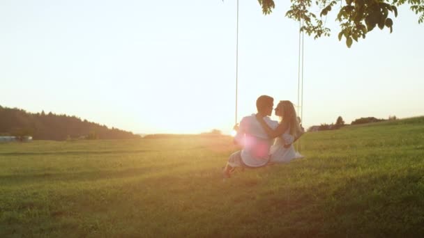 スローモーション 夕日に満たされた田舎でスイングキスに新進のガールフレンドとボーイフレンドを抱きしめた 自然の中で夏のデートにカップルの絆 抱擁し 揺れるロープのスイングにキスを共有 — ストック動画