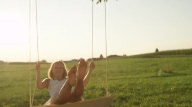 Slow Motion Close Up Portrait: Doğada yaz güneş ışınları salıncak gibi parlayan altın saçlı mutlu genç kadın. Beyaz bir sundress gülen bayan bahar kırsalında eğlenceli yalınayak sallanan