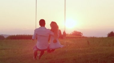 Slow Motion: Aşık çift sıcak bir yaz akşamı ağacın altında bir salıncak kucakladı. Erkek arkadaşı güneşli doğada sallanan salıncak takılan gülümseyen kız arkadaşı. Mutlu gençler salıncakta gülüyor.