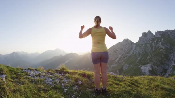スローモーションハッピー女性は 黄金の夕日に岩の山頂で勝利を収め 彼女の腕を上げます 日の出時に山の頂上に到達したときに腕を伸ばすハイキング女の子 日々の目標を達成する若いトレッカー — ストック動画