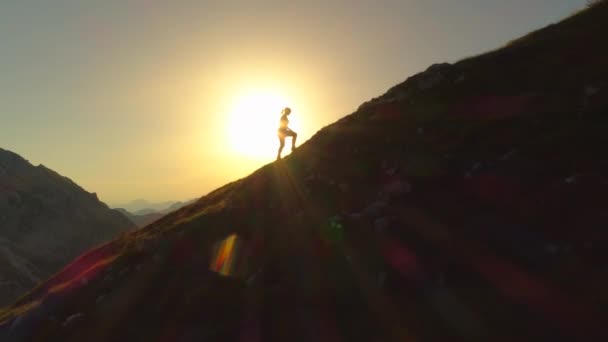 空中シルエット 彼女は頂上に向かう途中で傾斜地形を登るように若い女性ハイカーの上を飛ぶ 日没前に山頂に到達しようとする女性 美しい景色の中で上り坂を登る女性 — ストック動画