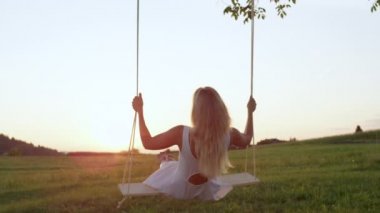 SLOW MOTION CLOSE UP: Lonely young blonde woman in white dress swinging under tree watching the sunset. Girl wrapped up in thought shakes her head and observes the sunset by herself on a rope swing.