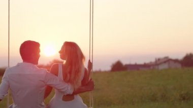 SLOW MOTION CLOSE UP: Joyous girlfriend and boyfriend kissing and laughing on swing on summer evening in countryside. Young couple enjoying last sunrays on romantic date whilst swaying on rope swing.