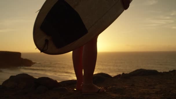 慢动作特写无法辨认的冲孩站在岩石峭壁上凝视着太阳落山时的海洋 冲浪后 年轻的运动员欣赏美丽的风景 有冲浪板看日落的女人 — 图库视频影像