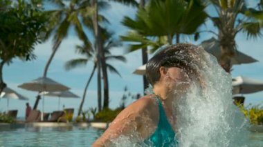 Slow Motion, Close Up: Turkuaz bikinili kadın, uzun kahverengi saçlarıyla camsı havuz suyu püskürtüyor. Havuzda rahatlatıcı dalış üzerinde oyuncu kadın gezgin başını batırır ve ters baş duymak çevirir.