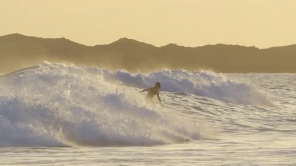 スローモーション 黄色の夕日が自然を照らし 波に乗ってサーファー 楽しい海の波に乗ってアクティブな休暇を楽しむ男性観光客 カラフルな自然の中で波に乗るサーファーの映画撮影 — ストック動画